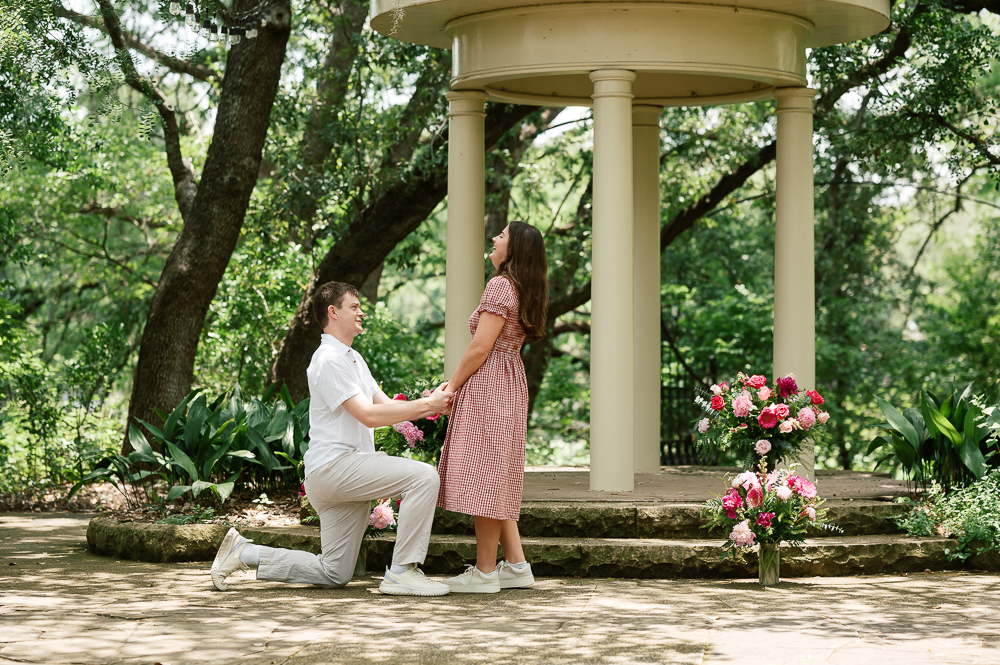 a man in a white shirt and tan pants is on one knee proposing to a girl who is laughing in front of the Temple of Love at Laguna Gloria in Austin. They are surrounded by lush greenery and beautiful large hot pink floral arrangements