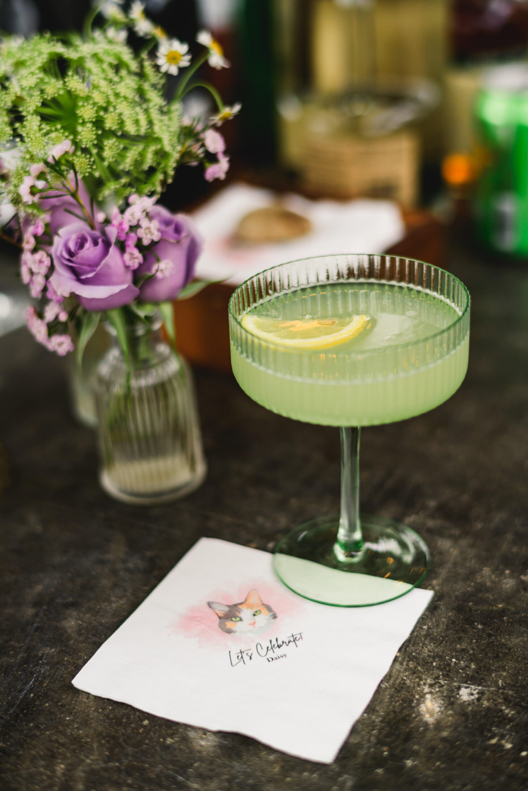 wedding cocktail in green coupe glass resting on a custom cocktail napkin of a cat that says "let's celebrate"