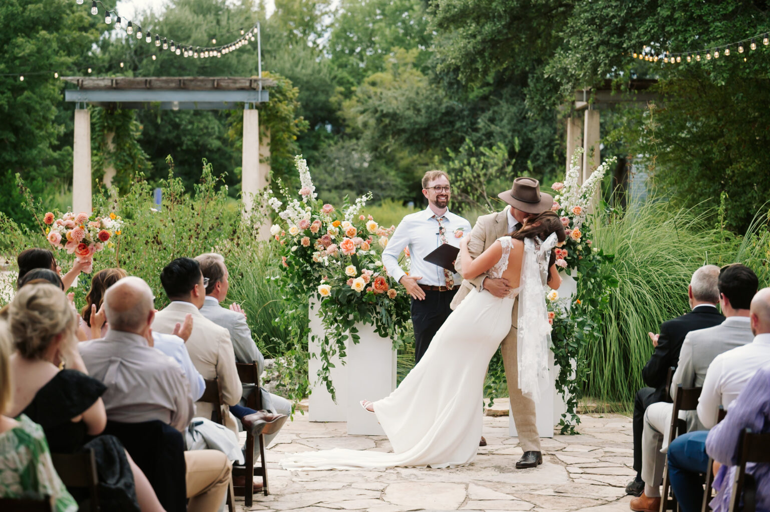 A couple shares a romantic kiss during their outdoor wedding ceremony at the Wildflower Center in Austin. The bride is in a white dress, and the groom in a tan suit and hat. Guests watch, seated among lush greenery and flower arrangements, under string lights.