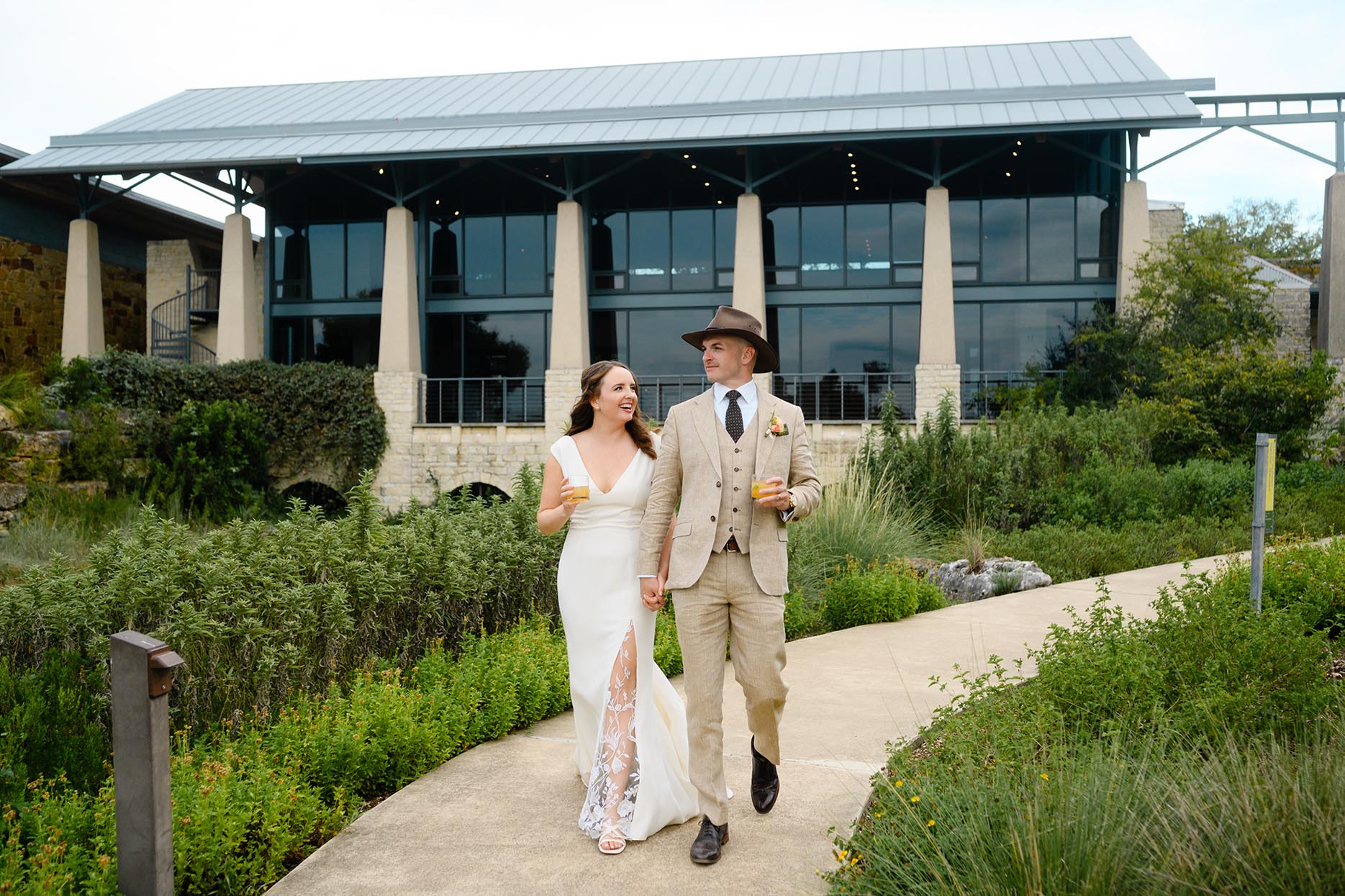 A bride in a white dress and a groom in a tan three-piece suit walk together with cocktails during portraits at the Ladybird Johnson Wildflower Center