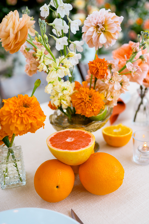 A sunny table setting with vibrant orange and yellow flowers in vases, ripe oranges, a halved grapefruit, and a lit tea candle, creating a cheerful atmosphere.