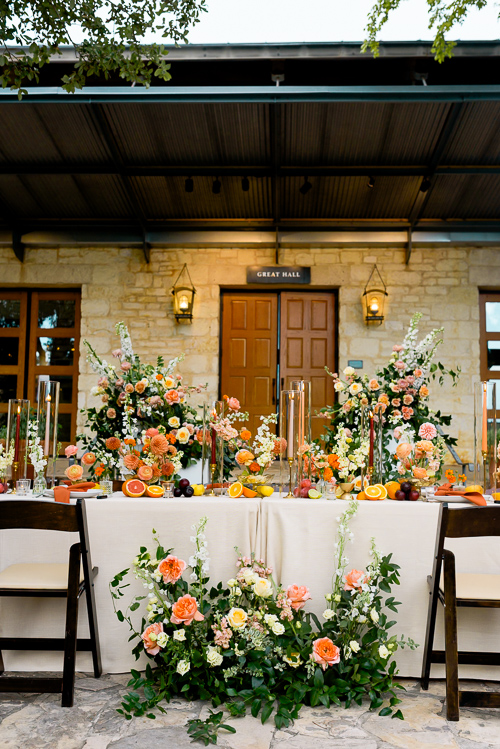 Outdoor dining set up with a long table adorned with vibrant orange and peach floral arrangements and sliced fruits in the courtyard at the Wildflower Center. Elegant and festive summertime wedding ambiance.