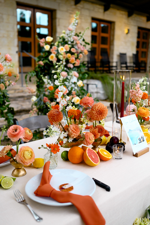 Elegant outdoor table setting with orange and peach flowers, citrus fruits, and a knotted orange napkin on a white plate, exuding a vibrant, festive summer wedding vibe.