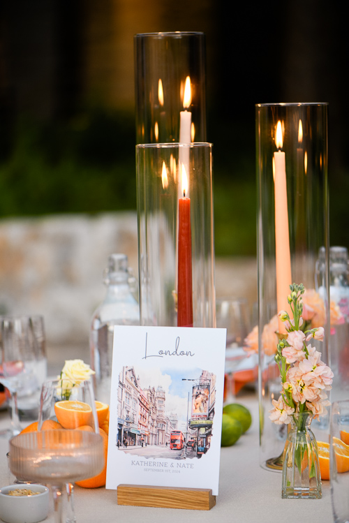 Tall glass candles illuminate a table with a card reading "London" featuring city sketches. Bright flowers and fruits add a vibrant touch. 