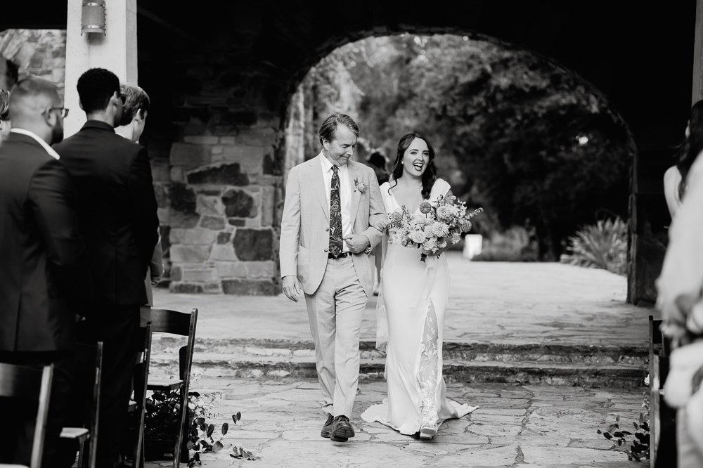 A black-and-white photo of a bride walking down an aisle at the Ladybird Johnson Wildflower Center with a man in a suit. She is smiling and holding a bouquet, surrounded by guests seated on either side.